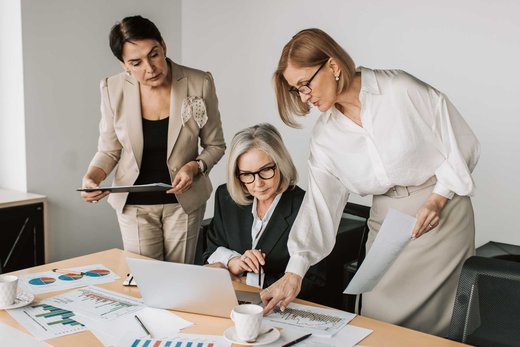 Three colleagues reviewing printed charts and data around a laptop at a conference table