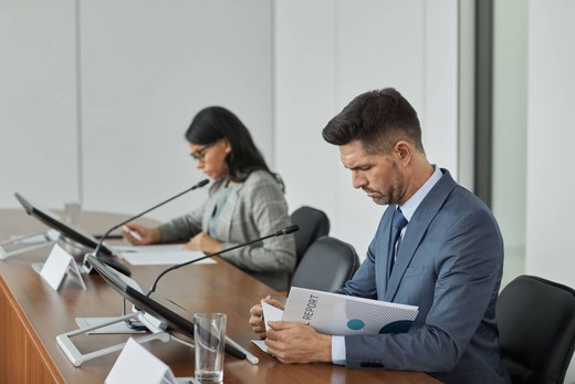 A man in a suit reviews a printed report at a conference table while a colleague reads documents beside him, microphones and water glasses on the desk