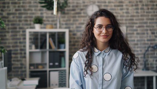 Smiling professional woman with curly hair and glasses standing in a bright, modern office
