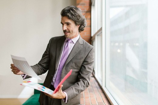 A man in a suit reviews a printed report and a color-coded folder beside a tall office window