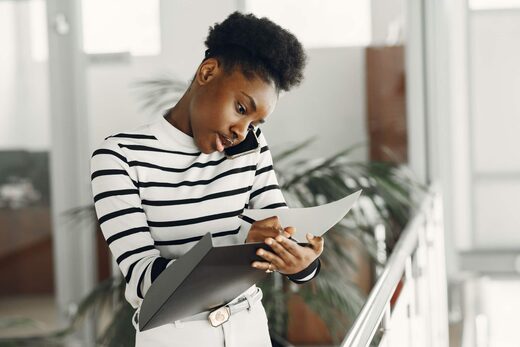 A professional woman takes notes on a clipboard while on a phone call in a bright, plant-filled office atrium