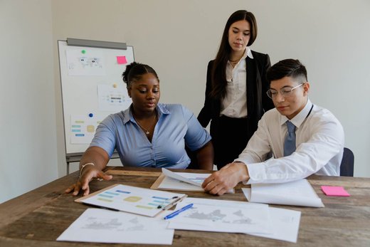 Three colleagues review printed charts at a table, with a flowchart-covered whiteboard behind them