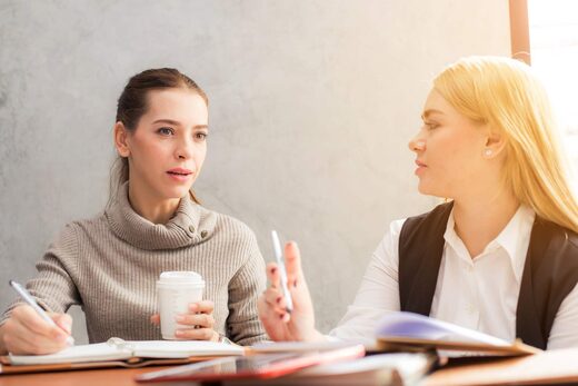 Two women in business casual attire talk across a sunlit table, one holding a coffee cup and taking notes