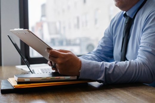 Professional reviewing a clipboard document beside an open laptop, city buildings visible through the window