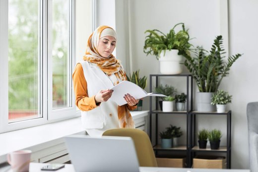 Woman in a hijab reading a document in a bright office with plants in the background