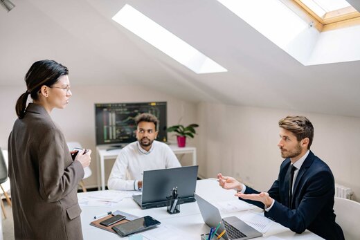 Three colleagues in a bright skylit office discussing data on laptops around a table