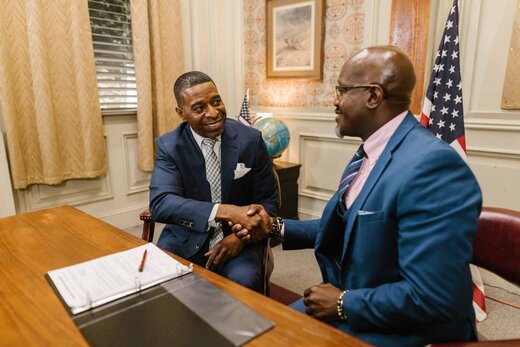 Two executives in navy suits shaking hands across a desk in a government-style office with an American flag