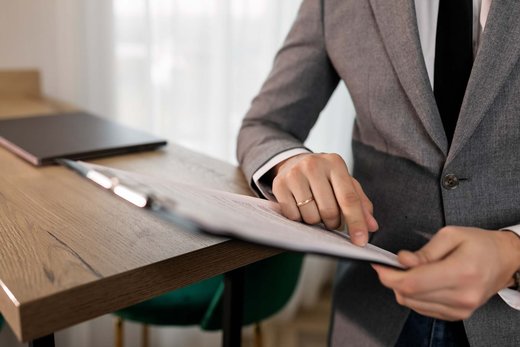 A man in a gray blazer reviews a signed report on a clipboard at a wooden desk