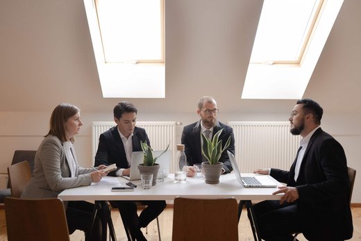 Four colleagues in business attire meeting at a table beneath bright skylights, two working on laptops