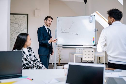 A presenter in a suit points to a hand-drawn upward trend chart on a whiteboard while two colleagues look on in a bright office