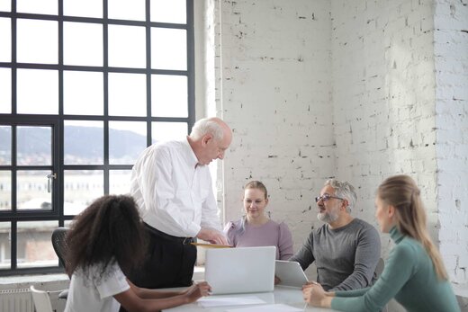 Senior leader briefing a seated team around a laptop in a bright, loft-style office
