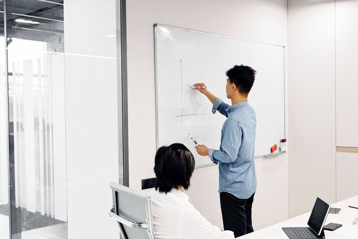 A man sketches an axis diagram on a whiteboard while a colleague looks on, with a tablet on the desk beside them