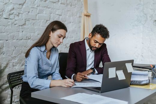 Two colleagues reviewing printed notes and a laptop together at a desk against a white brick wall