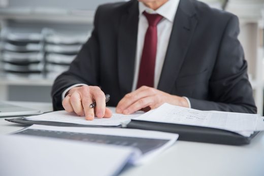 Businessman in a suit reviewing a data table on paper alongside an open laptop at a desk