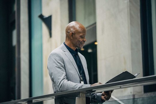 A senior man in a grey blazer smiles while reading an open black folder at a glass railing inside a marble office atrium