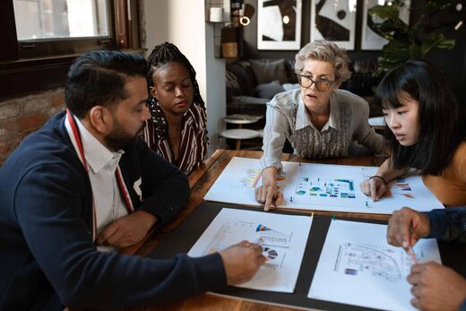 Five colleagues review printed charts and diagrams around a wooden table in a bright loft office with exposed brick and framed art