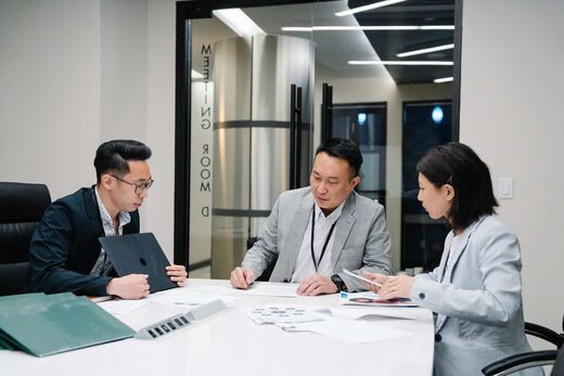 Three colleagues in dark blazers reviewing documents together around a table in a bright meeting room