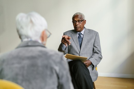 Senior consultant in a grey suit gesturing while explaining a point to a colleague, gold folder in hand
