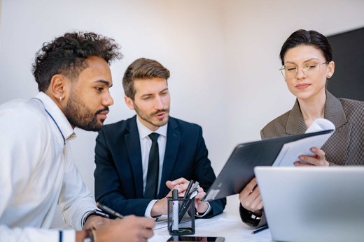 Three colleagues in business attire reviewing a document and laptops together in a bright white office