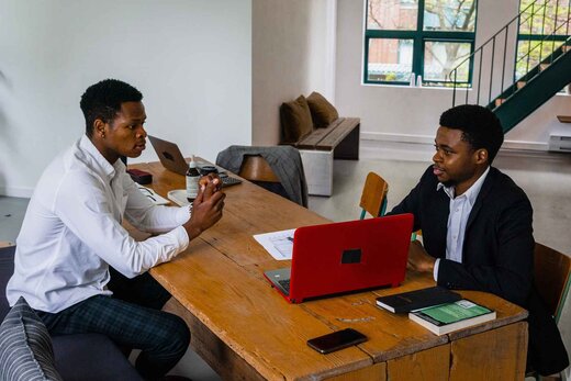 Two colleagues at a shared table reviewing documents on a laptop, one explaining a point to the other in a bright loft office