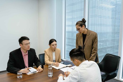 Four colleagues in a bright glass-walled meeting room reviewing documents and a tablet together, one standing to point out a detail