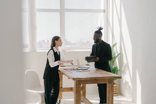 Two colleagues in conversation at a wooden table in a bright office, city skyline visible through the windows behind them