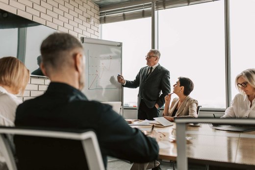 A senior leader in a grey suit presenting a hand-drawn trend chart on a flipchart to seated colleagues in a bright, window-lit conference room