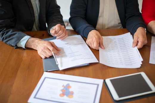 Two people's hands reviewing and annotating multi-page application forms on a wooden table