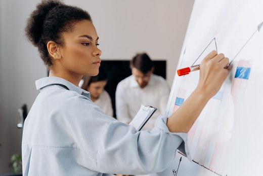 A woman sketches a chart on a whiteboard while two colleagues confer in the background of a bright office