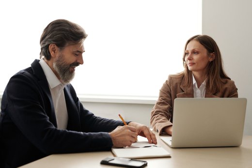 A bearded advisor taking notes across a desk from a colleague working on a laptop in a bright office