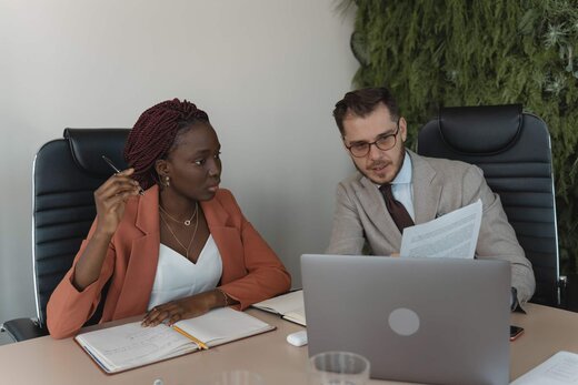 A woman in a coral blazer holding a pen looks over at a colleague in a beige suit pointing at a laptop screen, in a meeting room with a living plant wall