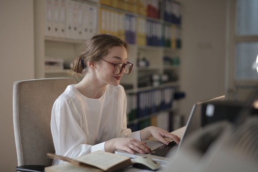 A regulatory professional reviews documentation on a laptop at a desk in a bright office, with binders of records on the shelving behind her.
