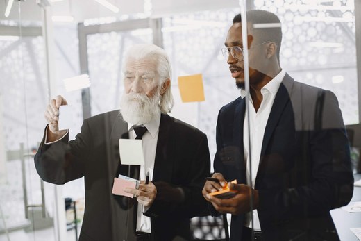 An older consultant and a younger colleague reviewing sticky notes together through a glass wall