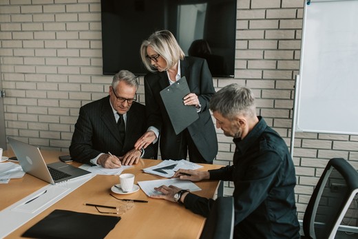 Two colleagues in dark suits reviewing and signing documents at a boardroom table while a third stands over them holding a clipboard