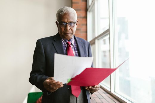 A senior executive in a red tie reviews documents from a red folder beside a bright window in an office with an exposed brick wall