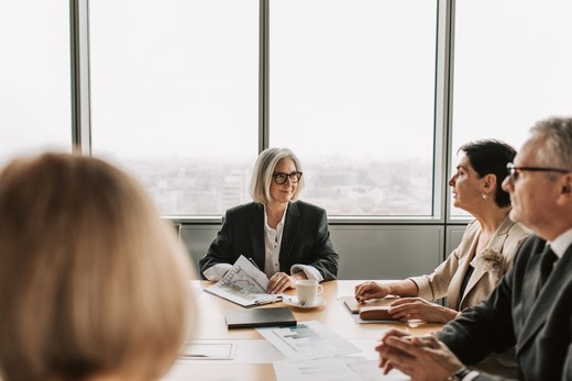 A senior consultant leads a discussion around a conference table with colleagues reviewing printed charts, a city skyline visible through floor-to-ceiling windows.