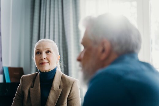 Senior regulatory advisor in a beige blazer and blue turtleneck listening closely to a colleague in soft window light