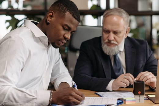 A senior advisor and colleague reviewing a document together at a desk, one hand on a stamp