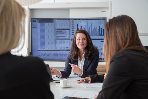 A woman in a navy blazer discussing data displayed on a large boardroom screen with two colleagues, papers and coffee cups on the table between them