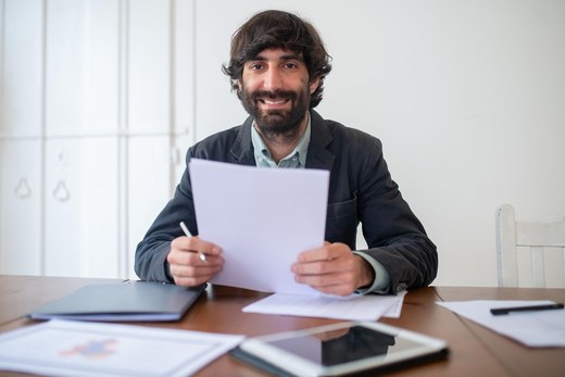 A smiling man in a blazer reviews a paper document at a wooden desk covered with folders, printed charts, and a tablet