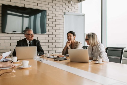 Three senior colleagues smiling in conversation around a boardroom table with laptops and coffee cups, in a bright brick-walled conference room with floor-to-ceiling windows