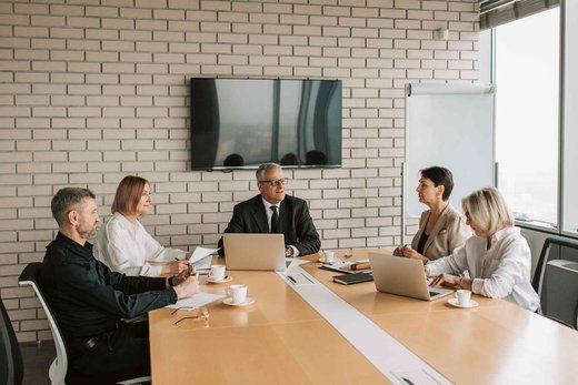 Two colleagues in business attire discussing documents at a wooden conference table with a monitor behind them
