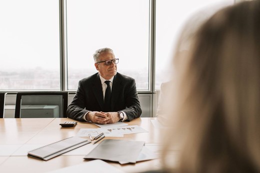 Senior consultant in a dark suit sitting at a boardroom table, listening thoughtfully with a city skyline visible through floor-to-ceiling windows