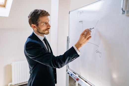A man in a navy suit sketches a plan and rising timeline curve on a whiteboard in a sunlit office