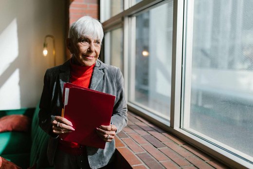 Senior regulatory professional in a grey blazer holding a red folder beside a bright office window