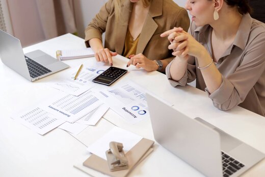 Two colleagues in blazers review printed reports and a calculator at a bright office desk