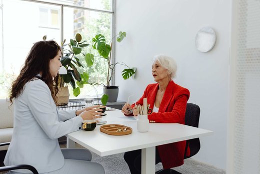 Two women in blazers talking across a table in a bright office with plants