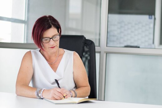 Senior regulatory professional with red hair and glasses writing notes at a desk in a bright glass-walled office