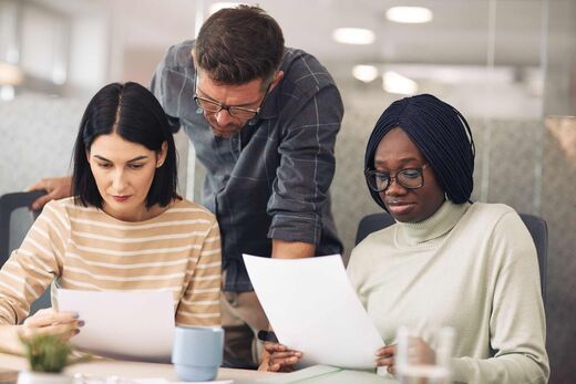 Three colleagues reviewing printed documents together in a glass-walled office