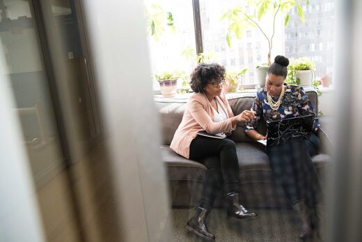Two professional women review notes together on a sofa in a sunlit high-rise office, with a city skyline visible through the window behind them